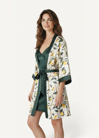 A woman wears a white satin robe with lemon and bird print green trim and belt in a green floral nightgown and robe set