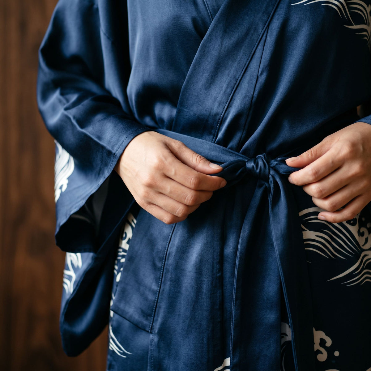 Close-up of hands tying the sash of an indigo satin kimono robe with a white wave print